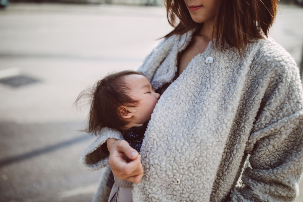 Pretty young mom carrying a sleeping baby with the baby carrier in the street - Latest Fashion & Lifestyle Trends What Is Breastfeeding?